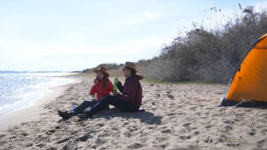 İki kadın sahilde oturuyor, biri kovboy şapkası takıyor. Turistler kızarmış sosis yer ve bira içerler. Deniz kenarında aktif bir hafta sonu. Lezbiyenlerin göl kenarında randevusu var.