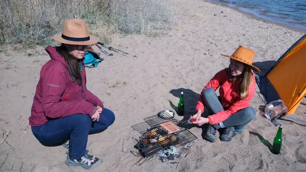 İki kız arkadaş sahilde oturuyor ve kamp ateşinin üstünde ızgarada sosis kızartıyorlar. Deniz kıyısında piknik. Çadırlı turistler hafta sonlarını aktif olarak geçiriyorlar.