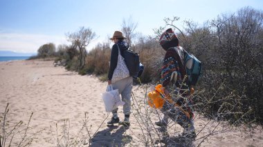 İki kişi sırt çantaları, çantalar ve sarılı bir çadırla sahil boyunca yürüyorlar. Arkadaşlar okyanus kenarında piknik yapmak için toplandılar. İnsanlar sahilde sakin bir gezintinin tadını çıkarıyorlar.