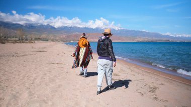 İki gezgin deniz kıyısında geziniyor. İki kişi plaj boyunca mavi bir gölün arka planında yürüyorlar. Seyahat, macera ve turizm