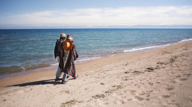 Mavi okyanusun arka planında sahilde gezinen bir çift. Arkadaşlar güneşli bir günde plaj boyunca dolaşıyorlar. Turizm, yürüyüş ve maceralar. Denizde tatil.