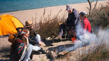 Arkadaşlar sahilde balık kızartıyorlar. Bir grup insan deniz kenarında bir kamp ateşinin etrafında oturuyor. Kamp ateşi tütüyor, insanlar sohbet ediyor. Turistler temiz havada dinlenir.
