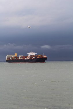 Büyük bir kargo gemisi fırtınalı hava ve koyu gri gökyüzü ile denizde yol alıyor.