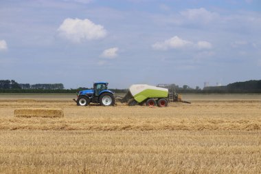 Büyük bir balyası olan bir traktörle saman tarlası ve yazın Hollanda kırsalında bulutlu mavi bir gökyüzü.