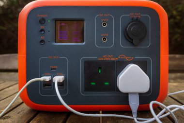 Close up of portable power station solar electricity generator outdoors on wooden table with electronic devices wires and cables plugged in. Wireless charging lithium battery backup for use off grid.
