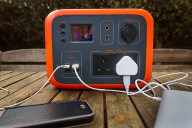 Portable power station solar electricity generator outdoors on wooden table with mobile phone and laptop computer electronic devices charging. Wireless charging lithium battery backup for use off grid