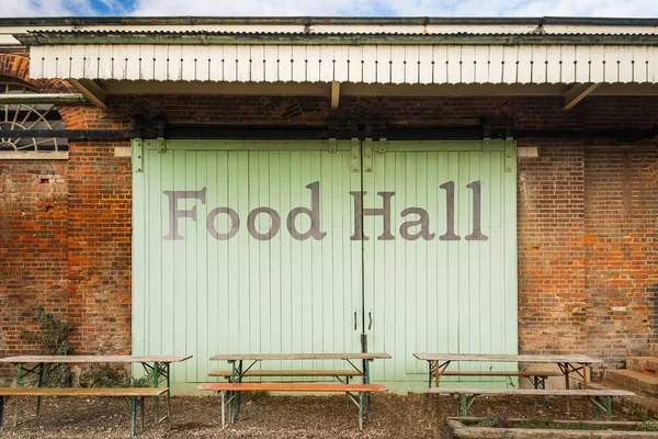 Exterior of rustic food hall building with large wooden green barn doors, red brick walls and benches in front. Food Hall words painted on doors.