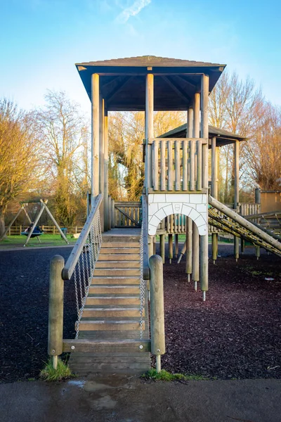 Childrens playground wooden play structure fort with wood step ladder bridge and high tower. Daytime outdoor image with blue sky, trees in background and soft, safe floor covering. Portrait image.