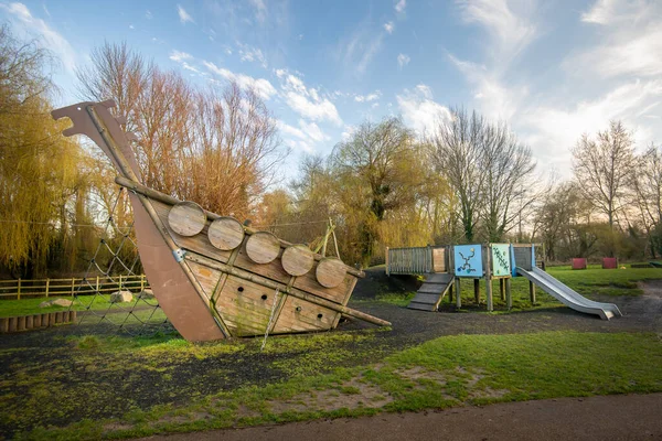 Wooden boat climbing frame structure in outdoor natural public playground with slide in background, grass, trees and safe soft floor covering surface. Blue sky, daytime scene. Idyllic childhood.