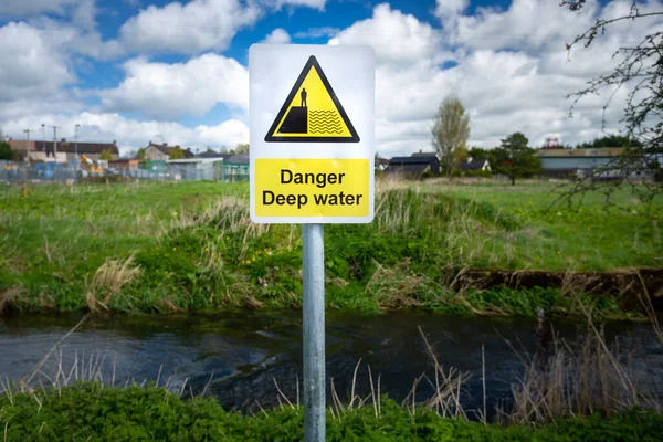 Yellow, black Danger Deep Water sign on rural river bank. Green field, buildings in far background. Hazard signage alerting public to dangerous deep water. Risk of falling in and drowning. Slip hazard
