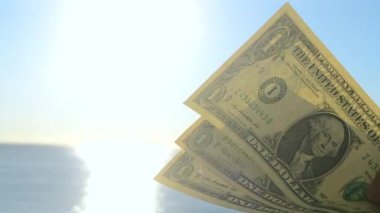 Elinde dolar banknotları tutan kişi. Parlak güneş, mavi gökyüzü ve durgun mavi denizin arka planında bir dolar değerinde. Bir dolarlık banknotlar mavi deniz gökyüzünün zemininde