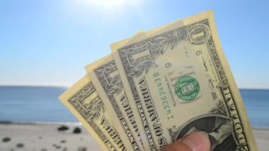 Elinde bir dolarlık banknotlar tutan bir adam denizin arka planına, kumlu kumsala ve güneşli yaz gününde mavi gökyüzüne karşı. Para, finans, para, seyahat, turizm, tatil, dinlenme kavramı