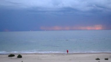 Kırmızı ceketli yaşlı bir adam sabah koşusunu deniz kıyısında kumlu bir sahilde yapıyor. Şafak vakti mavi fırtına bulutlarının, yağmurun ve mavi denizin zeminine karşı..