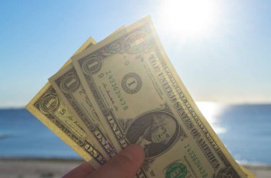 Elinde dolar banknotları tutan kişi. Parlak güneş, mavi gökyüzü ve durgun mavi denizin arka planında bir dolar değerinde. Bir dolarlık banknotlar mavi deniz gökyüzünün zemininde