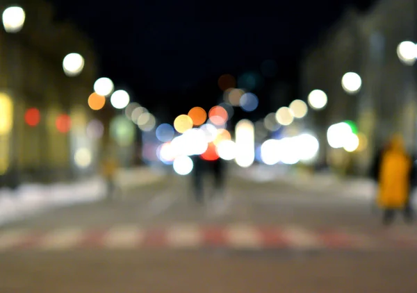Blurred background. Night city. Blurred silhouette of buildings, bokeh spots of glowing lanterns, blurred silhouettes of people walking on the street in the evening. City square, black night sky