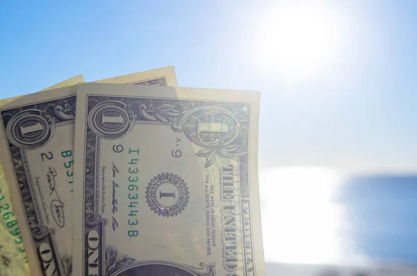 stock image Person holding in hand paper bills of dollars with face value of one dollar on background of brightly shining sun, blue sky and calm blue sea. One-dollar paper banknotes on backdrop of blue sea sky