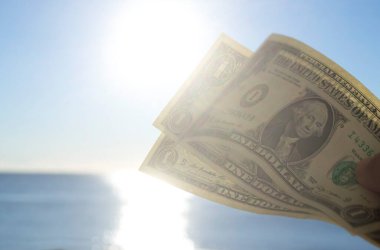 Elinde dolar banknotları tutan kişi. Parlak güneş, mavi gökyüzü ve durgun mavi denizin arka planında bir dolar değerinde. Bir dolarlık banknotlar mavi deniz gökyüzünün zemininde
