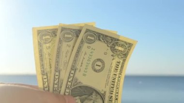 Elinde dolar banknotları tutan kişi. Parlak güneş, mavi gökyüzü ve durgun mavi denizin arka planında bir dolar değerinde. Bir dolarlık banknotlar mavi deniz gökyüzünün zemininde