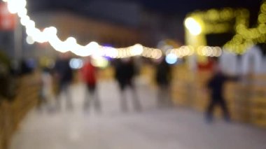 Arka plan bulanık. Birçok insan kış akşamı şehirde açık havada buz pateni pistinde kayıyor, parlak tatil çelenkleri takıyor. Noel Yeni Yıl kutlamaları Tatil eğlence arkaplanı