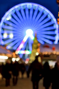 Dönme dolap mavi aydınlatma, Noel ağacı, yürüyen insanların siyah siluetleri, kış gecelerinde şehir binaları. Noel Kasabası. Güzel yeni yıl tatili geçmişi bulanıklaştırıyor