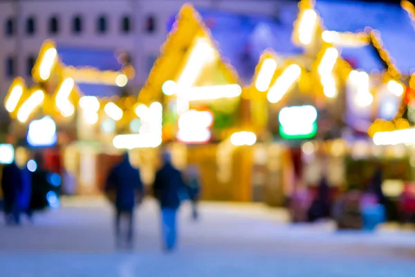 Blurred background. People walk in city square on winter night. Black silhouettes of people walking near houses decorated luminous illumination. White light bokeh blur spots from glowing house lights