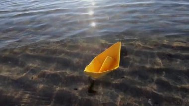 Deniz dalgalarında sallanan, şeffaf temiz su ve güneşli bir günde kumlu zeminde yüzen küçük sarı kağıt tekne. Origami gemisi yaklaşıyor. Hedef kavramı, seyahat, turizm hayal kurma seyahati