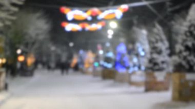 Bulanık arkaplan. Kış gecelerinde, yoğun kar yağışında, şehirde parlak ışıklar ve çelenklerle süslenmiş, süslenmiş Noel ağaçlarının arasında yürüyen insanlar.