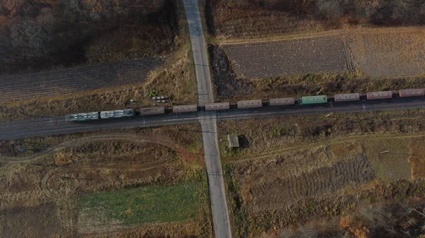 Top View Freight Train Passing Railway Crossing. Aerial Drone View Flight Over Intersection of Railway Tracks and Asphalt Road. Transportation, Rail freight, Delivery. Railway Wagon Rides on Railroad.