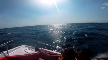Güneşli bir yaz gününde insanların süratle yelken açtığı beyaz bir motorlu tekne. Sürat teknesi deniz dalgalarında zıplıyor. Yat motorlu teknesi hızlı denize yelken açıyor. Motorlu tekne ile seyahat eden turistler. Güneş suyun üzerinde parlıyor