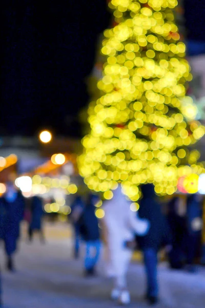 stock image Blurred background. Christmas tree, decorated glowing lights, building, blurry silhouettes of people walking city square at winter night. Beautiful New Year and Christmas holiday blurred background