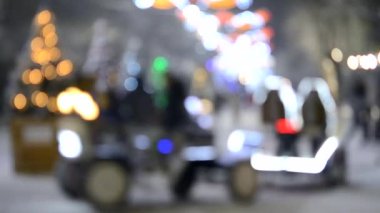 Arka plan bulanık. Işıklarla süslenmiş, parlak çelenklerle süslenmiş, büyük motosikletli, şehir meydanında, kış gecesi kar yağarken insanlara binen süslü Noel ağaçları.