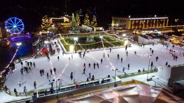 Birçok insan açık havada buz pateni pistinde paten kayıyor. Yılbaşı süsleri, süslemeler, kış gecelerinde parlak çelenkler. Yeni yıl Noel kutlaması. Havadan