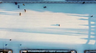 People skating on an open-air ice skating rink in the city in the evening during sunset in winter. Top view. People skating on ice rink. Long shadows of people on surface of ice. View from above