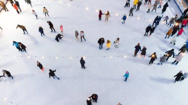 Kışın açık havada buz pateni pistinde paten kayan birçok insan görülüyor. Buz pateni manzarası. Şehir Parkı Buz Pisti. Kışın açık hava aktiviteleri. Tatillerde paten kaymak. Yeni yıl Noel Aydınlatmaları