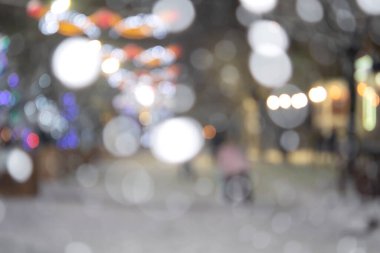 Bulanık arkaplan. Kış gecelerinde, yoğun kar yağışında, şehirde parlak ışıklar ve çelenklerle süslenmiş, süslenmiş Noel ağaçlarının arasında yürüyen insanlar.