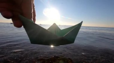 Adam kağıt tekneyi denizin yüzeyine koyuyor ve şafak vakti dalgaların üzerinde yüzüyor. Güneşli bir yaz gününde denizde yelken açan küçük bir kağıt tekne. Origami oyuncağı. Seyahat hedefi turizmi seyahat hayali olarak kabul et