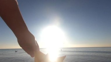 Parlak güneşli bir günde küçük kağıt tekneyi denizin yüzeyine koyan kadın eli. Origami gemisi deniz dalgaları üzerinde yelken açar. Parlak güneş, güneşli yol, aydınlık gökyüzü, sakin deniz. Rüya turizmi konsepti
