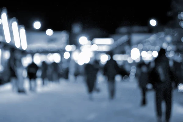 Blurred background. People walk in city square on winter night. Black silhouettes of people walking near houses decorated luminous illumination. White light bokeh spots glowing house lights Blue color