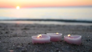 Three candles in shape of heart burn on sand on the sea beach near the sea waves against the background of the shining sun during the dawn of the sunset. Romantic romance mood. St. Valentines Day.