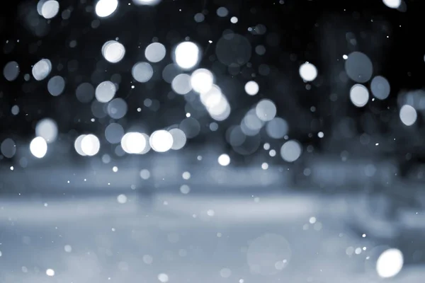 Blurred background. City view, lights, falling snow, night, street, bokeh spots of headlights of moving cars. Urban backdrop winter scenery of street in city night. Lantern light, snowfall. Blue color