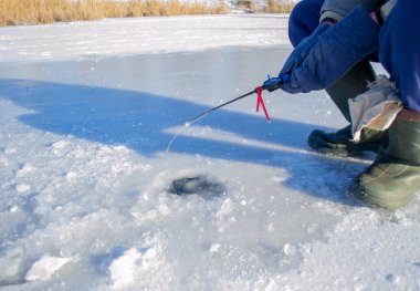 Winter ice fishing. Fisherman sitting near ice-hole and holding small fishing rod in hands. Ice fishing in winter. Small fishing rod stands near hole in the ice of river on sunny day. Winter activity