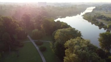 Landscape view green trees in park, calm river in city, multi-storey urban buildings, pink sun glare. Panoramic in summer morning. Urban landscape. Cityscape. Aerial drone view. Top view. Lifting up