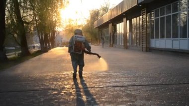 Man in airtight suit sprays disinfecting liquid a pavement on a street in the city against the backdrop of a sunset. Sanitation workers clean a crosswalk. Sanitary measures. coronavirus pandemic