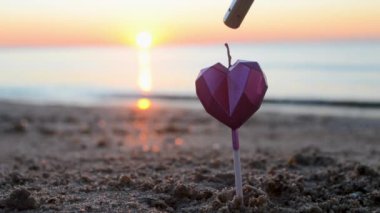 Heart shaped candle burning on sand of beach near sea waves during sunset dawn. Burning candle in form of heart in sand against backdrop of rising sun. Romantic romance mood. St. Valentines Day.