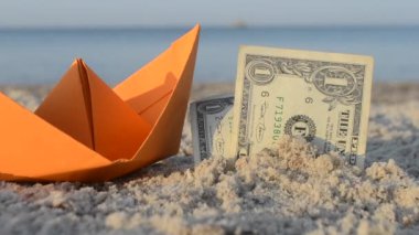 Turuncu kağıt tekne ve kumlu sahilde kumsalda kumsalda iki kağıt para. Arka planda mavi deniz, güneşli yaz gününde yakın plan. Finansman parası dolarları nakit seyahat turizmi tatili