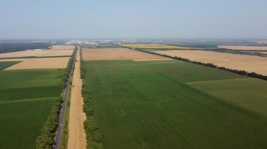 Farklı tarımsal yeşil bitkiler, olgun sarı buğdaylar, hareket halindeki arabaların ve şehrin bulunduğu güzel panoramik manzara. - Hava aracı. Tarım mahsulü manzarası