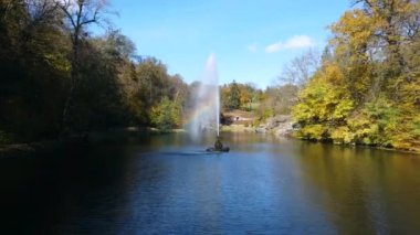 Fountain with rainbow in middle of lake between trees with yellow leaves in park on sunny autumn day. Decorative fountain in center of lake. Natural park arboretum in autumn. Natural landscape. Aerial