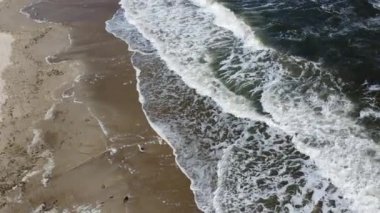 Yukarıdan gelen deniz dalgaları. Güneşli bir günde, deniz kıyısındaki kumlu sahilde beyaz köpüklü deniz dalgaları yuvarlanıyor. Kumsalın üzerinde uçuyor. Üst Manzara. Yukarıdan bak. Deniz manzaralı su manzarası. Hava aracı görünümü