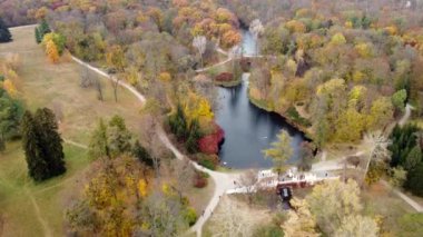 Beautiful scenery view of an autumn park with trees with yellow fallen leaves, lakes, architecture, glades and people walking along dirt paths on an autumn day. Flying over the autumn park. Top view.