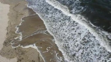Yukarıdan gelen deniz dalgaları. Güneşli bir günde, deniz kıyısındaki kumlu sahilde beyaz köpüklü deniz dalgaları yuvarlanıyor. Üst Manzara. Yukarıdan bak. Hava aracı görüntüsü. Kumsalın üzerinde uçuyor. Deniz Burnu su manzarası.